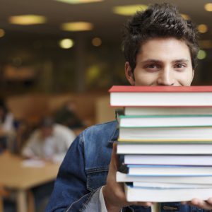 Student holding books in front of face in library, portrait